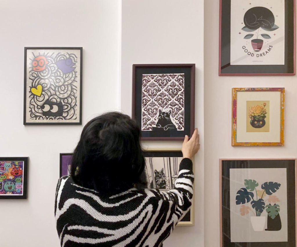 Woman holding a picture frame in front of a wall covered with framed cat artworks